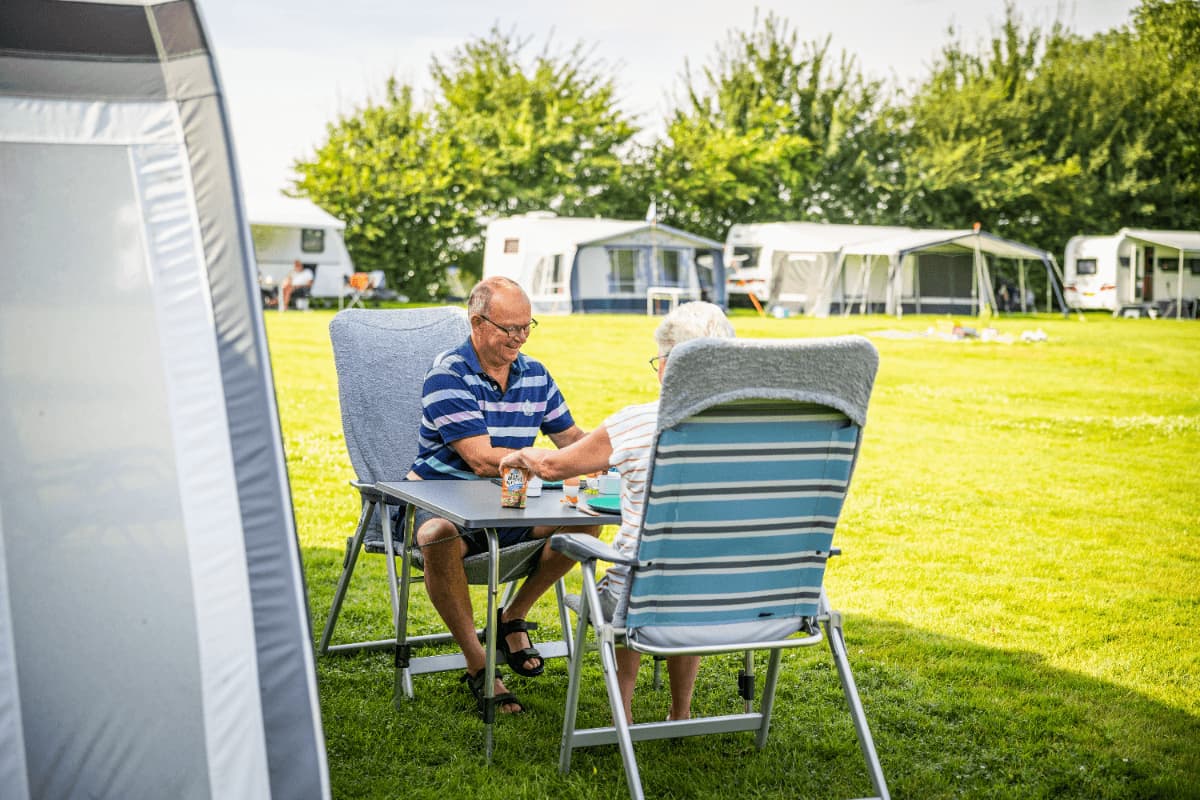 Op de camping zijn ruime staan plaatsen, die op de achtergrond te zien zijn. Twee mensen nemen een lekker ontbijt op hun campingstoelen voor de tent